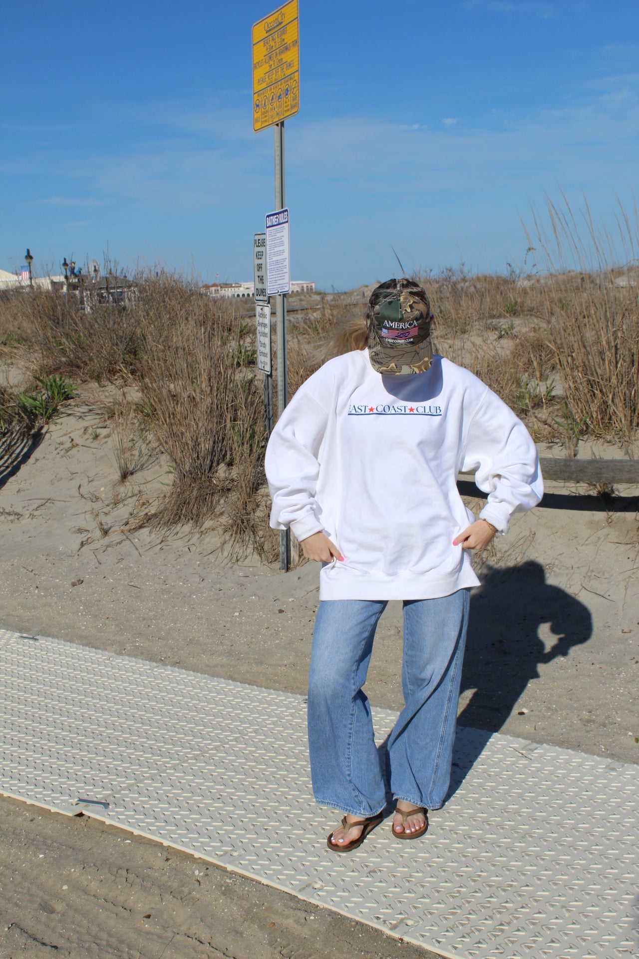 Person wearing a white sweatshirt with text, standing on a pathway near a beach.