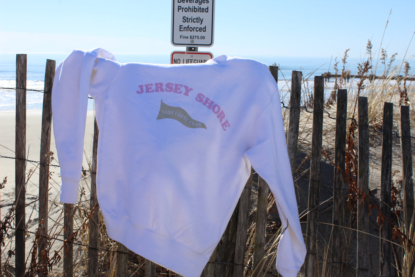 White sweatshirt with 'Jersey Shore' text on a beach fence with ocean view