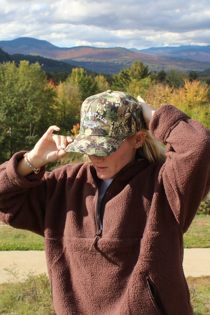 A woman outdoors in a mountain landscape wearing a brown fleece pullover, beige leggings, and a camouflage baseball cap, adjusting the brim of the cap.