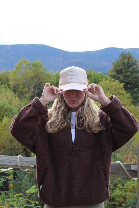 A woman outdoors wearing a dark brown fleece pullover and a beige baseball cap with 'EAST COAST CLUB' embroidered on the front, with mountains and trees in the background.