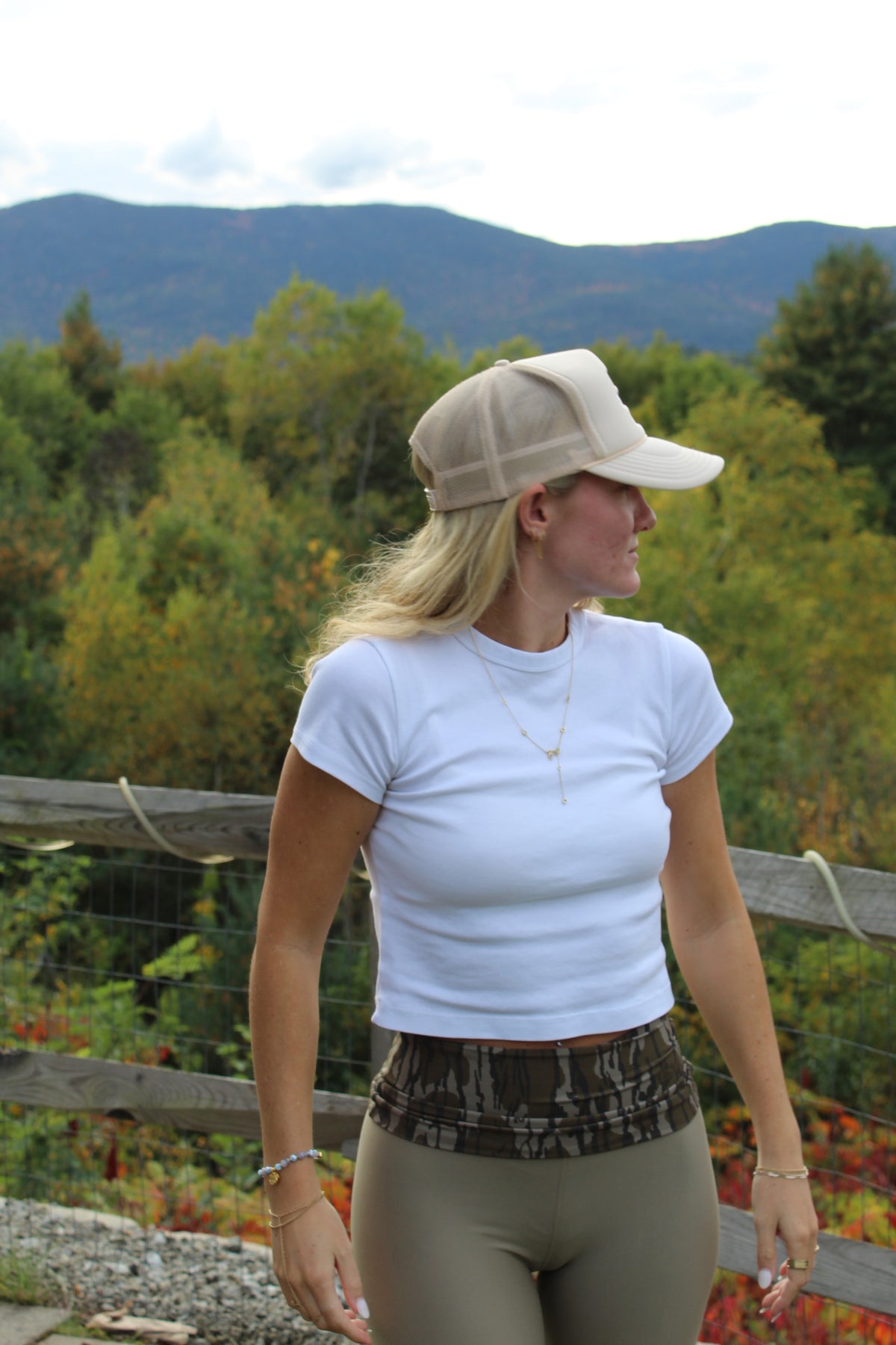 A woman wearing a white short-sleeve crop top, olive green leggings, a beige trucker cap, and a camouflage waistband stands outdoors with a wooden fence and forested mountains in the background.