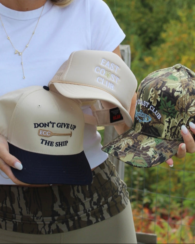 A woman outdoors holds three East Coast Club trucker hats: one beige with navy and embroidered text reading 'DON'T GIVE UP ECC THE SHIP', one beige with white embroidered text reading 'EAST COAST CLUB', and one camouflage pattern hat with white embroidered text reading 'EAST COAST CLUB'.