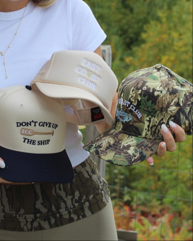 A woman outdoors holds three East Coast Club trucker hats: one beige with navy and embroidered text reading 'DON'T GIVE UP ECC THE SHIP', one beige with white embroidered text reading 'EAST COAST CLUB', and one camouflage pattern hat with white embroidered text reading 'EAST COAST CLUB'.