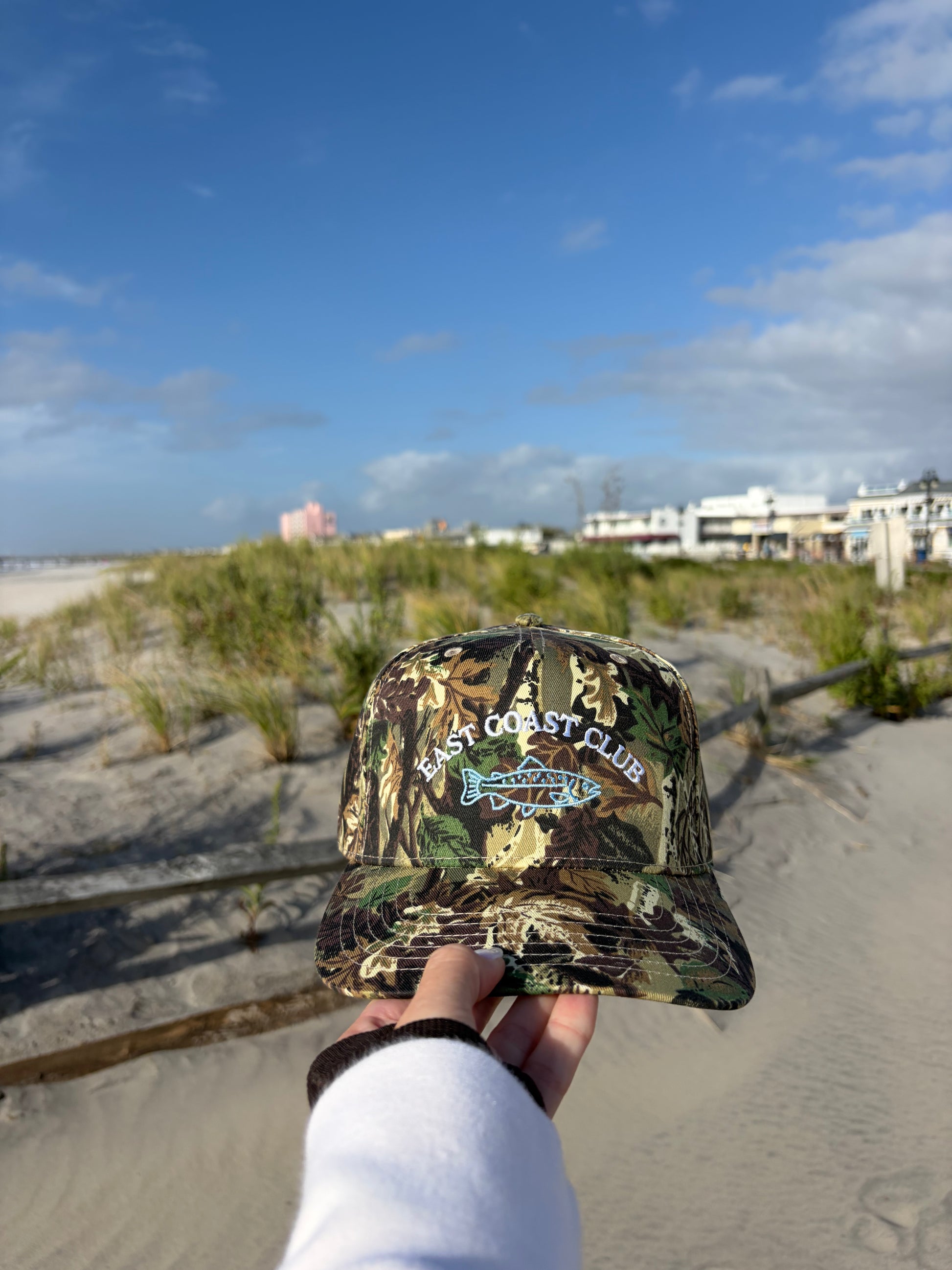 A camouflage baseball cap with 'East Coast Club' and 'Jaxx' embroidered on the front, held up by a hand against a beach dunes and blue sky background.