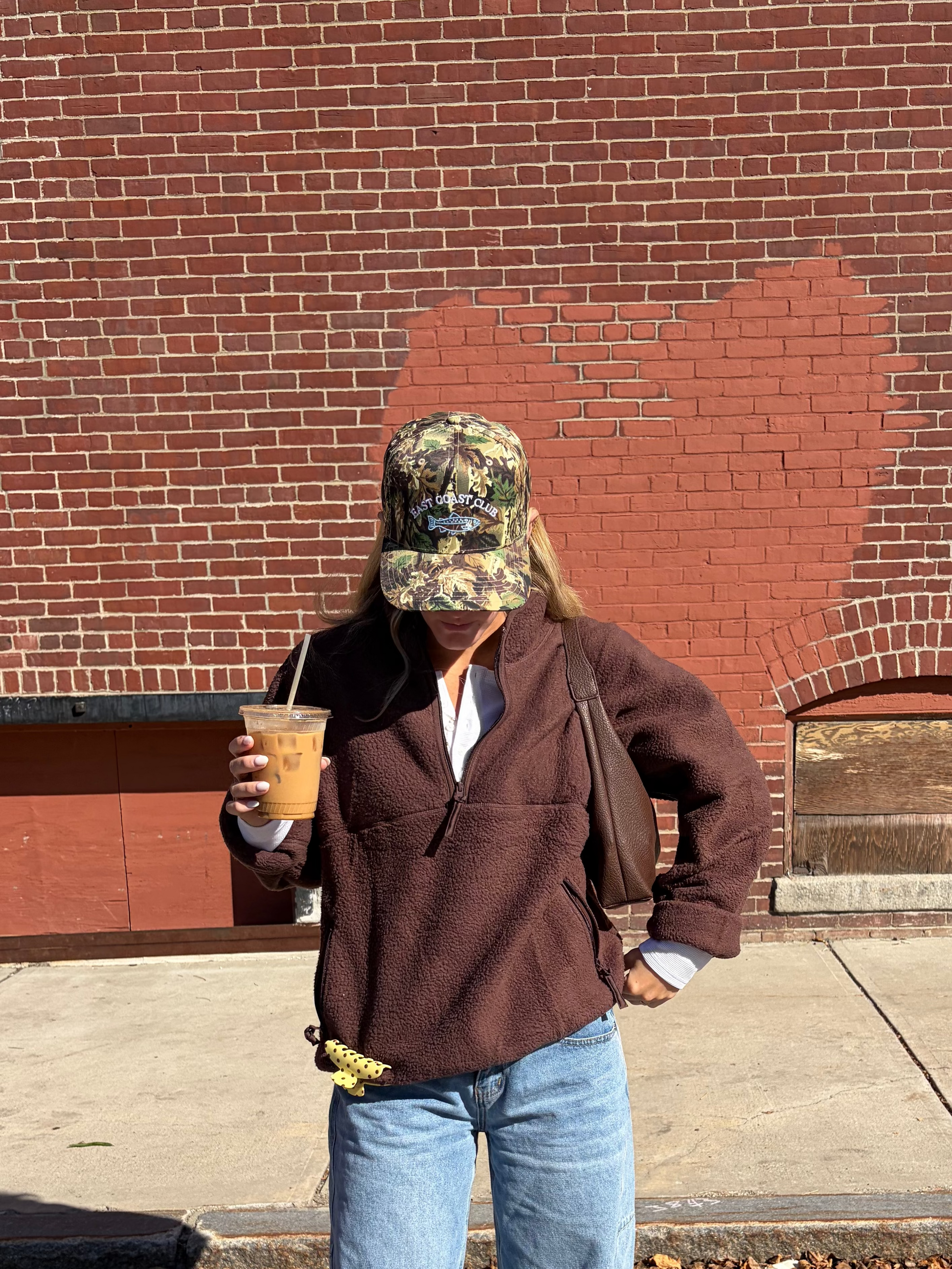 A person standing outdoors in front of a red brick building, wearing a brown quarter-zip fleece, light blue flared jeans, a camouflage baseball cap, and white sneakers, while holding an iced coffee.
